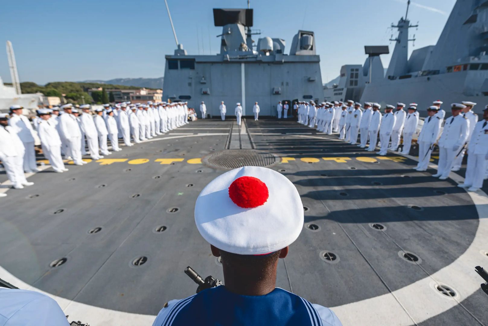 Une cérémonie militaire sur le pont d'un navire, avec l'équipage en tenue blanche de cérémonie. Au premier plan, un matelot avec coiffe à pompon au milieu des rangs.
