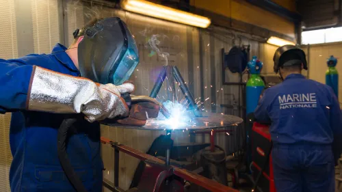 Un marin de spécialité opérateur industriel en atelier naval, en tenue bleu de travail, portant un masque à souder, en train de souder une pièce métallique