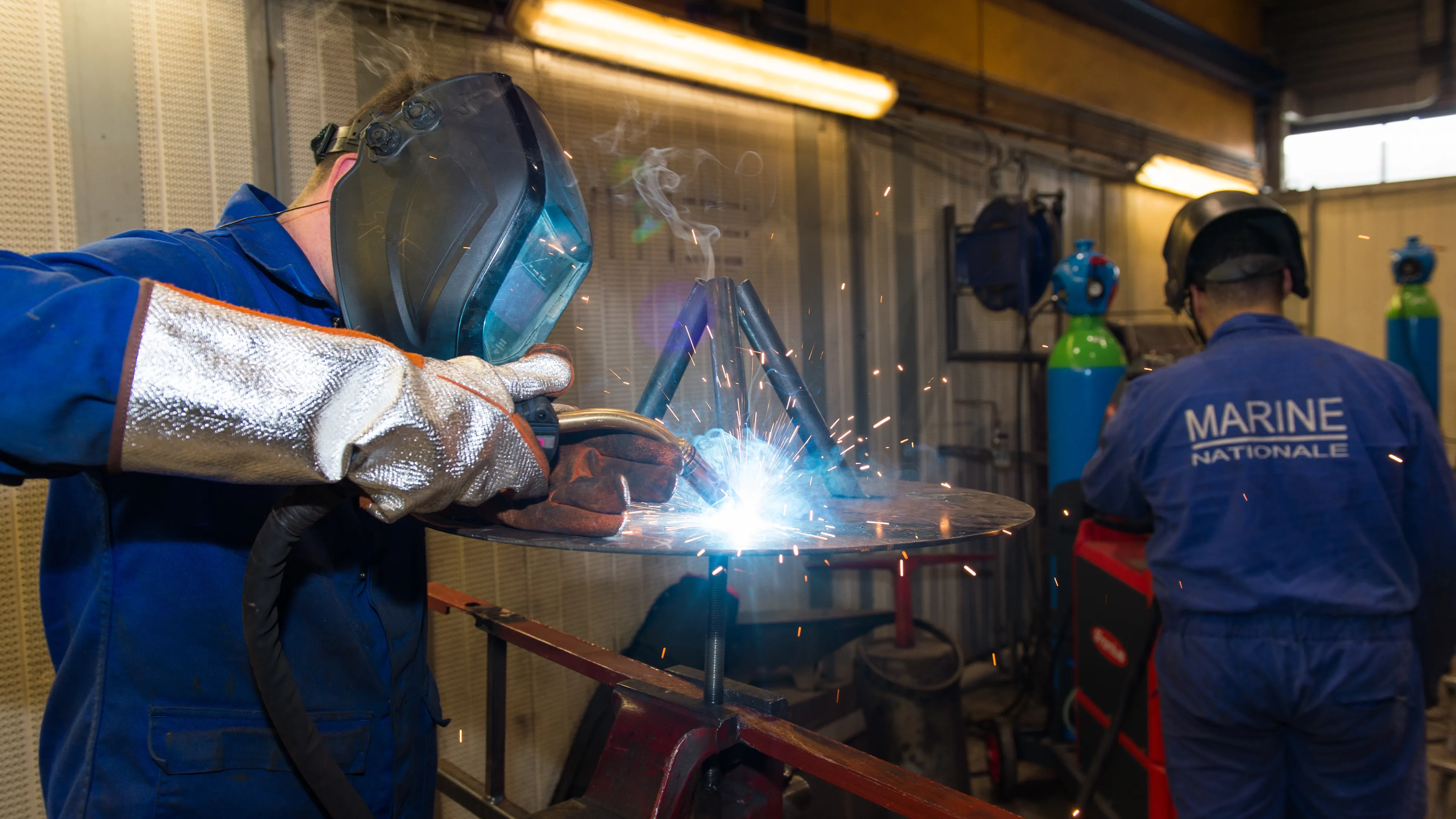 Un marin de spécialité opérateur industriel en atelier naval, en tenue bleu de travail, portant un masque à souder, en train de souder une pièce métallique