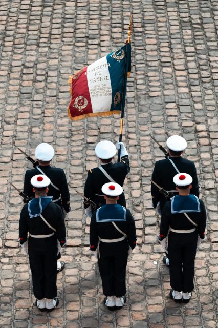 Un groupe de six marins alignés sur deux rangs pour une cérémonie. Tous en uniforme bleu, avec armes de cérémonie et drapeau Français.