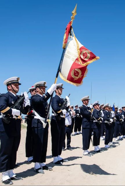 Des marins en rang, équipés d'armes de cérémonies et d'uniformes de cérémonie, sur une plage, avec le drapeau français.