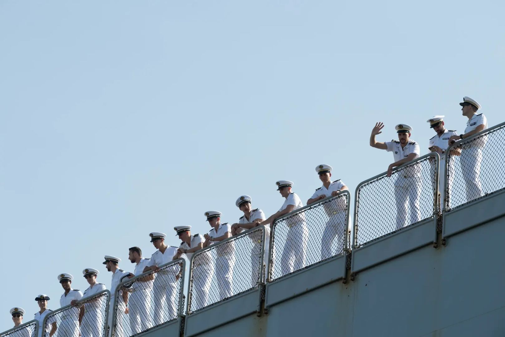 Des marins en uniforme blanc alignés sur le pont d'un navire, disant au revoir à leurs proches sur le quai.