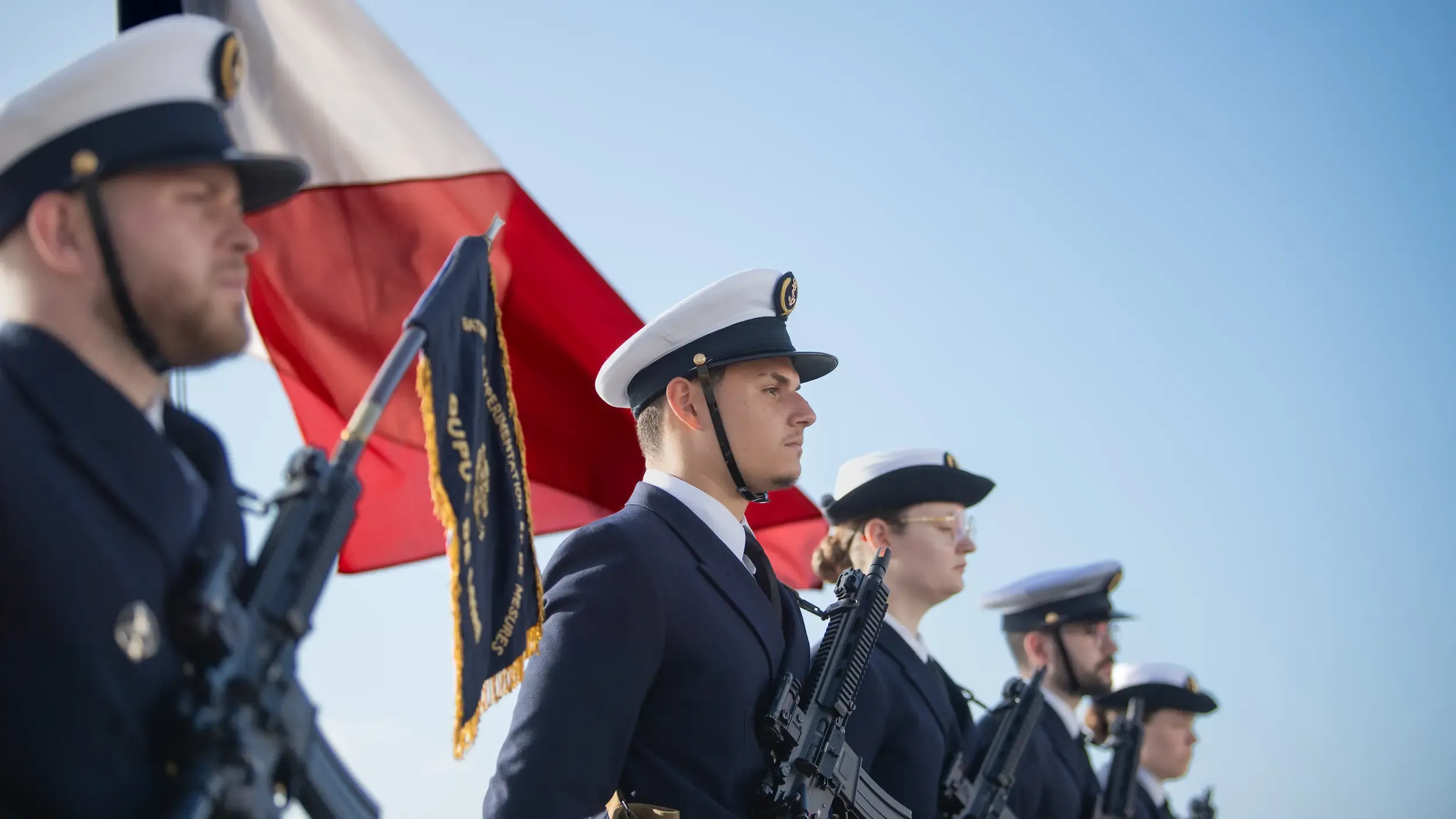 Un groupe de marins alignés en rang pour une cérémonie, uniforme bleu avec coiffe et armes, avec un drapeau français flottant au vent derrière eux.