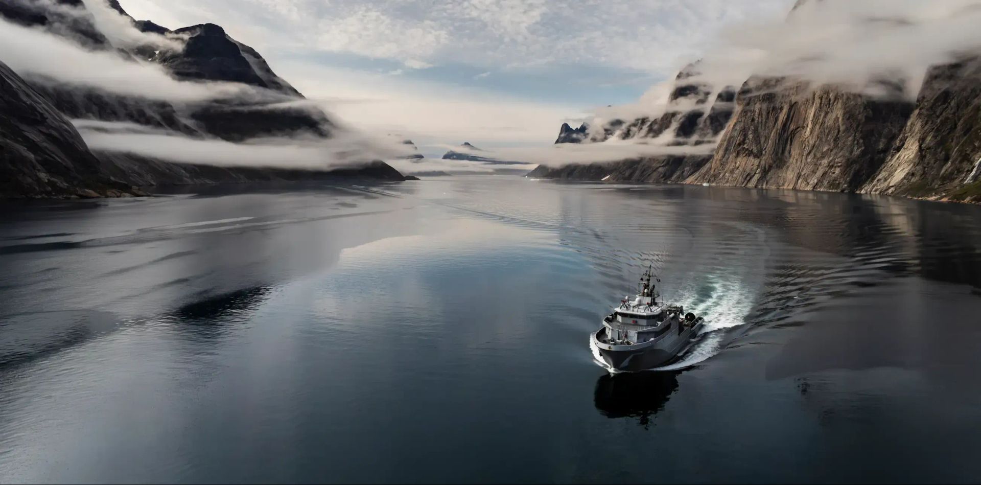 Un bateau de la marine nationale en train de naviguer des les eaux d'un fjord, l'eau est calme et il y a du brouillard sur les rochers environnant.