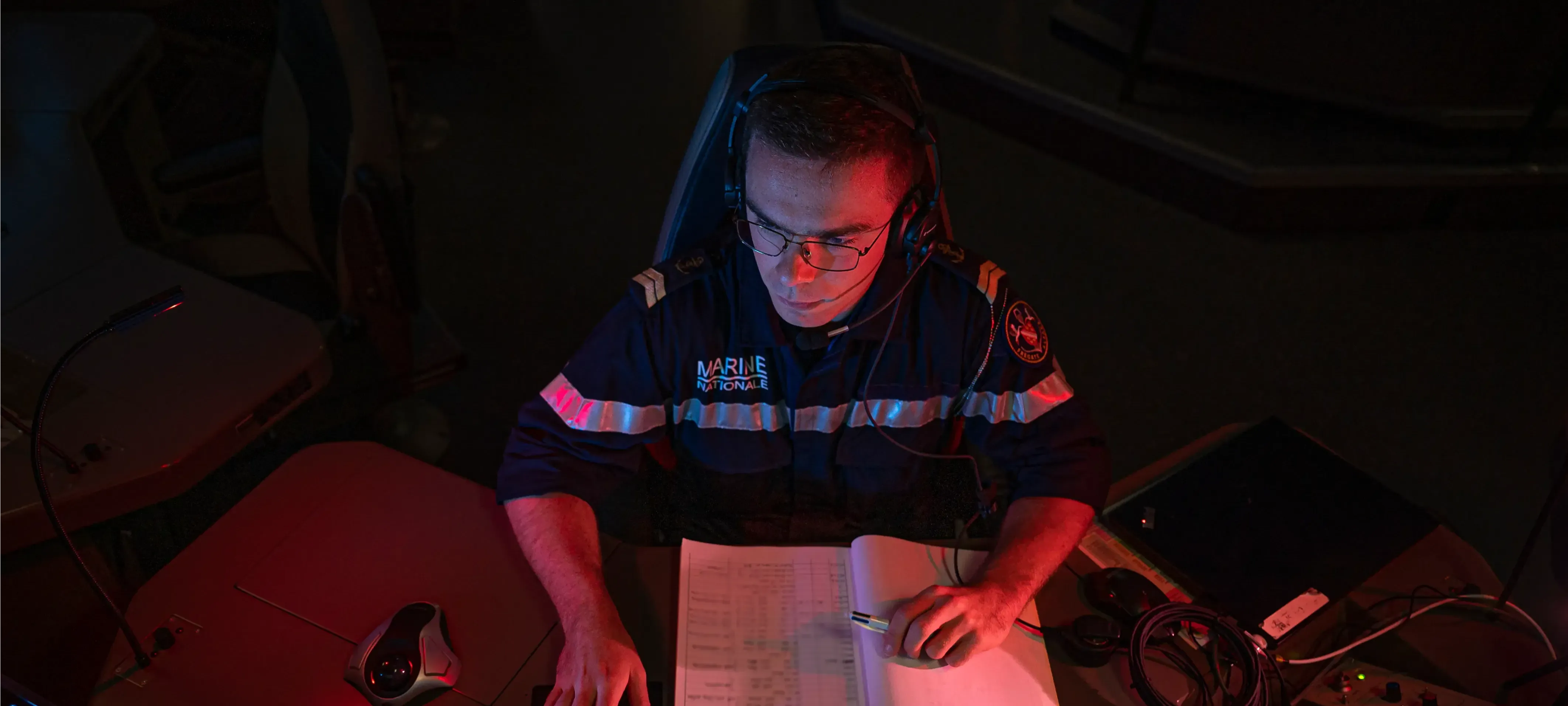 Un marin détecteur anti sous-marins au poste de travail pour analyser des données, s'aidant d'un carnet et d'un ordinateur