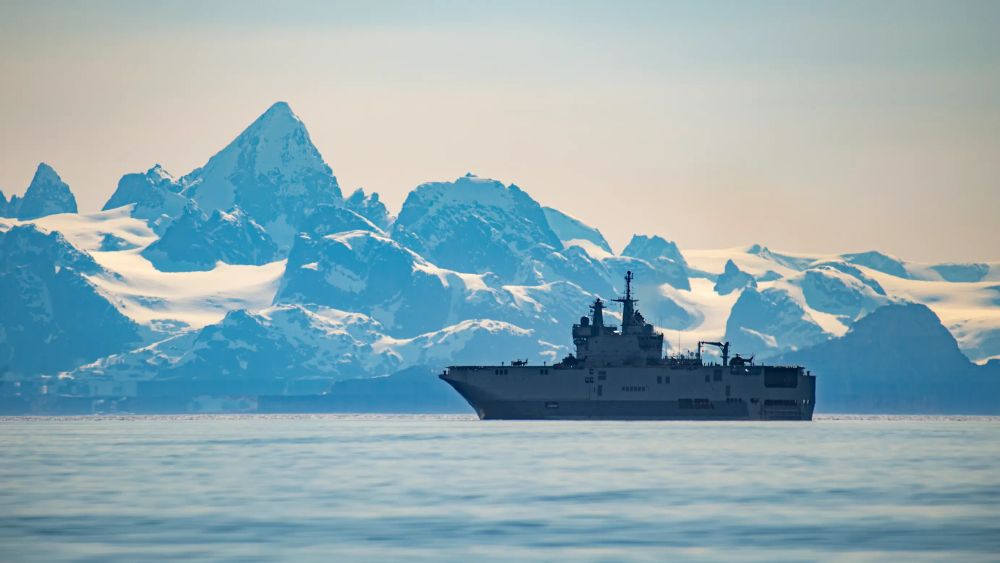 Un porte-hélicoptère amphibien en train de naviguer sur les mers du groenland, avec des montagnes enneigées en fond.