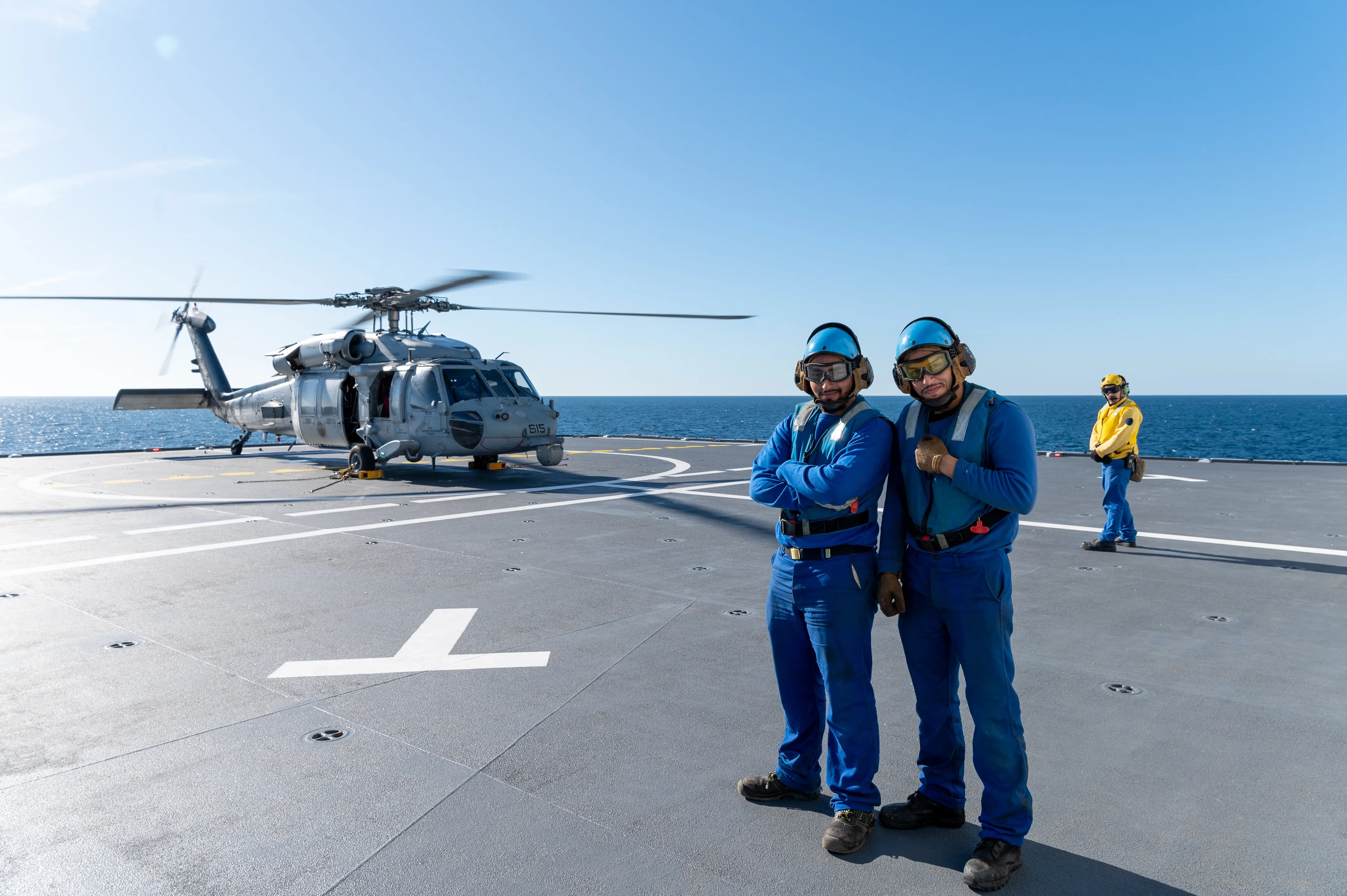 Deux opérateurs de manutention des aéronefs sur un pont avec en second plan un hélicoptère de la Marine Nationale