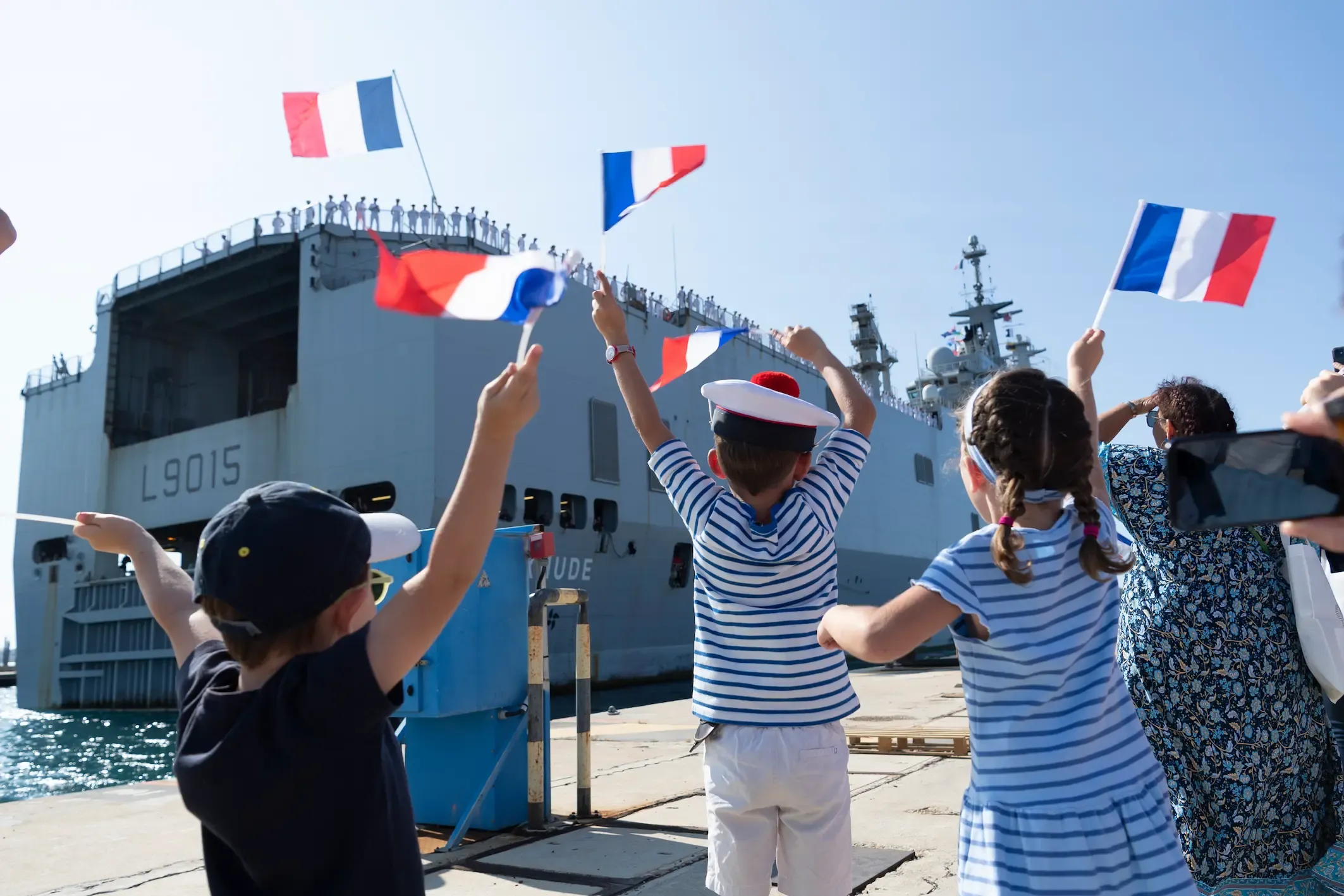 Des enfants de marins accueillant leurs parents de retour de mission, ils agitent des drapeaux français et attendent sur le quai que le navire accoste.