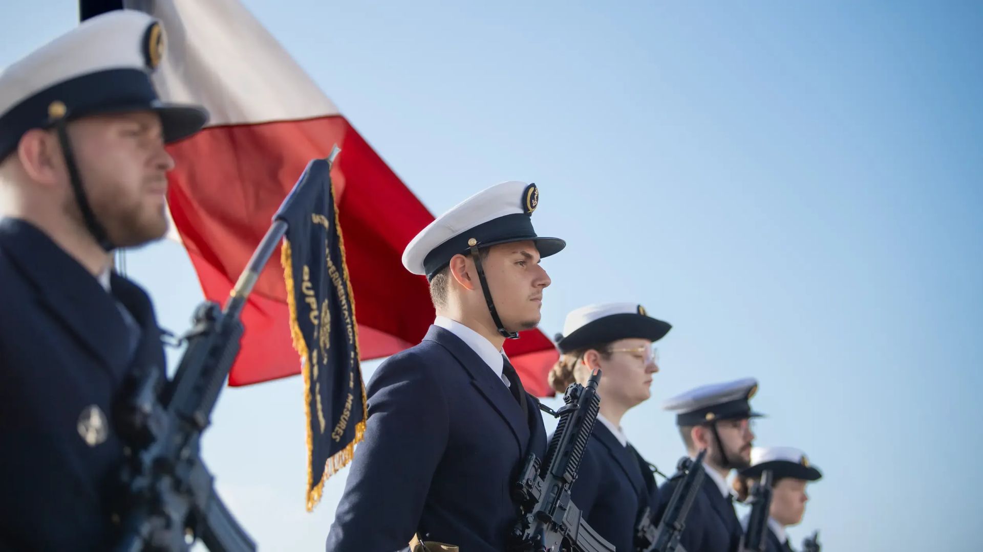 Un groupe de marins alignés en rang pour une cérémonie, uniforme bleu avec coiffe et armes, avec un drapeau français flottant au vent derrière eux.