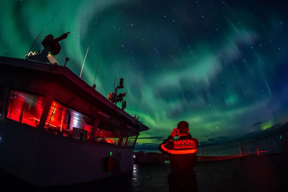 Un bateau de la marine nationale en mode nuit, avec un marin en train d'observer les aurores boréales vertes et bleus au dessus de lui.