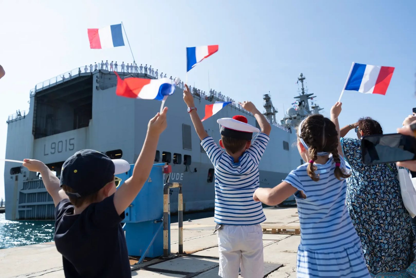 Des enfants de marins accueillant leurs parents de retour de mission, ils agitent des drapeaux français et attendent sur le quai que le navire accoste.