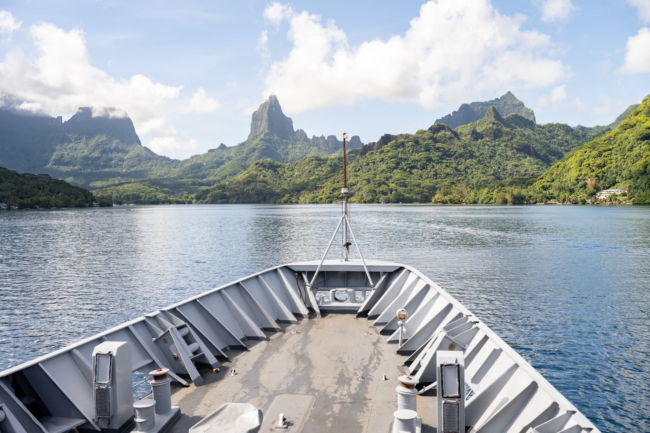 La photo représente la proue d'un navire de la marine française naviguant sur un baie paradisiaque entourée de montagnes et de végétation.