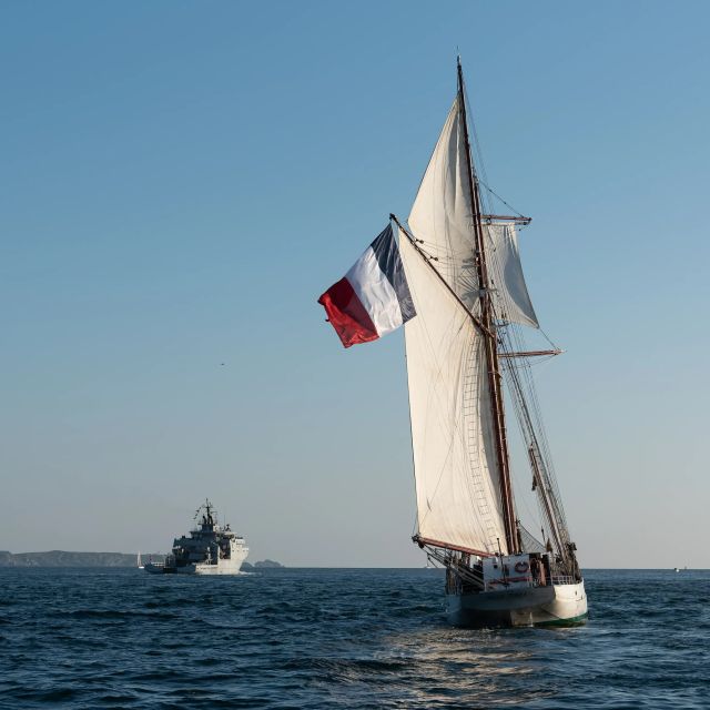 Un voilier de la marine avec pavillon français, naviguant sur une mer bleu, et un navire récent de la marine nationale en fond.